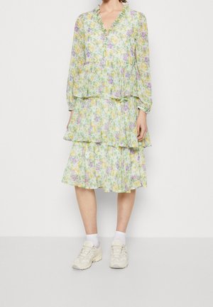 Robe florale à volants en vert clair avec des fleurs jaunes et violettes, dotée de manches longues et d'un col à volants. Associée à des chaussettes blanches et des baskets.
