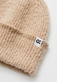 Knitted beige beanie hat with a textured pattern and a folded cuff, featuring a small white label with a black logo.