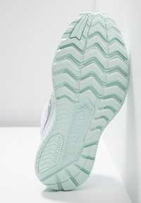 Bílá sportovní bota s texturovanou mátově zelenou gumovou podrážkou. Obsahuje cikcak vzor a vyvýšené prvky pro lepší přilnavost.
