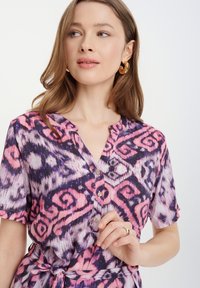 Knopfhemdkleid mit einem abstrakten Muster in Lila, Pink und Schwarz. Kurze Ärmel, Kragen und ein Bindegürtel in der Taille. Glatte Stofftextur.