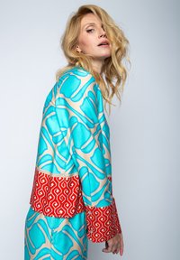 Kolorowa, wzorzysta jedwabna kurtka w kolorze aqua i beżowym z kontrastowym czerwonym geometrycznym pasem na dole oraz szerokimi mankietami.