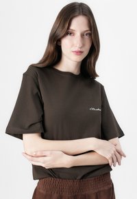 Bruine katoenen T-shirt met korte mouwen, voorzien van een eenvoudig wit geborduurd tekst op de voorkant. Zachte textuur en relaxed fit.