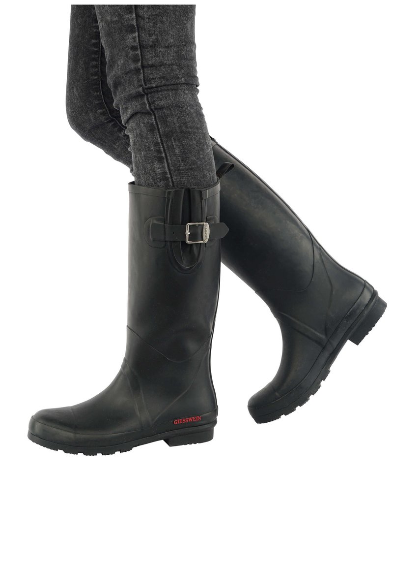 Bottes en caoutchouc noires jusqu'aux genoux avec une boucle, portées sur un jean foncé gris ajusté, jambes légèrement croisées, nom de la marque visible près de la semelle.