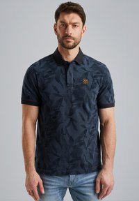 Polo en bleu marine avec un motif de feuilles, tissu texturé et col noir contrastant ; présente un petit logo noir et or sur la poitrine.