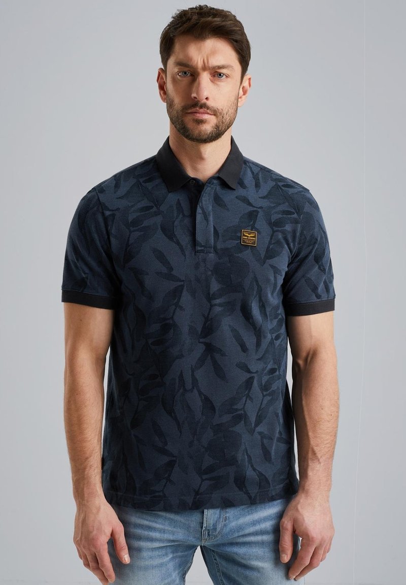 Polo en bleu marine avec un motif de feuilles, tissu texturé et col noir contrastant ; présente un petit logo noir et or sur la poitrine.
