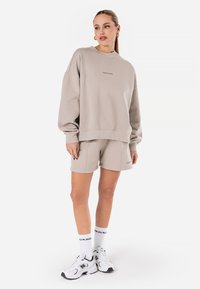 Beige oversized genser med ribbede mansjetter, sammen med matchende shorts. Hvite joggesko og sokker med logoaccents fullfører antrekket.