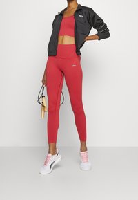 Top sportivo rosso a coste e leggings abbinati a una giacca nera. Scarpe sportive bianche e rosa. Tiene una corda per saltare.