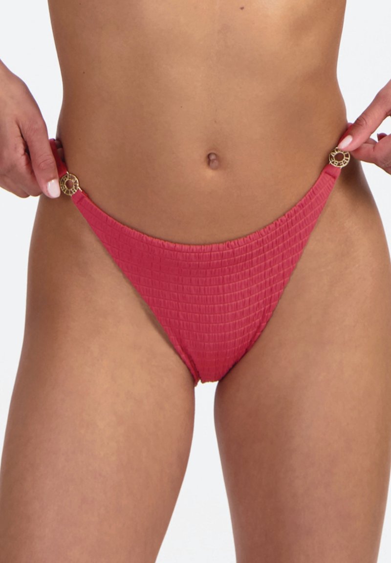 Rote Bikinihose mit Struktur und goldenen, runden Schnallendetails an der Hüfte, Hände richten die Seiten aus, vor einem schlichten hellen Hintergrund.