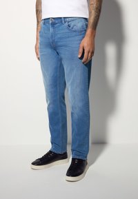 Jean en denim bleu clair avec une coupe droite, doté de détails de poche et d'une texture subtile, associé à des baskets noires.