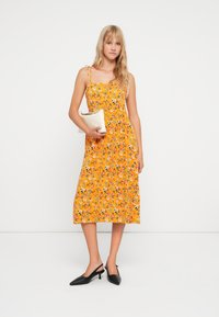 Vestido floral amarelo com um design fluido, alças finas e um decote com babados. Combinado com sapatos de salto preto tipo slingback e uma clutch branca.