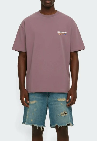 Man wearing a mauve oversized T-shirt with small "Barrow" logo on chest and distressed denim shorts with frayed hems.