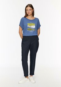 Niebieska koszulka z grafiką w kolorze zielonym, białym i czarnym, zestawiona z czarnymi zwężającymi się spodniami i białymi sneakersami. Minimalistyczny styl.