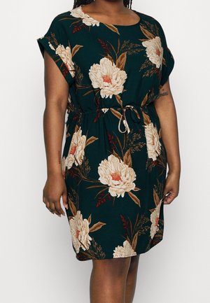 Kvinde iført en mørkegrøn kjole med store hvide og brune blomsterprint og snøre i taljen, står mod en ensfarvet baggrund.