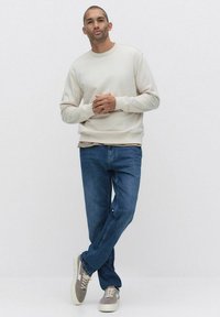 Jasno beżowa bluza z prążkowanymi mankietami, w połączeniu z niebieskimi dżinsami i szarymi trampkami. Model stoi w luźnej pozie, z rękami złożonymi. Tło neutralne.