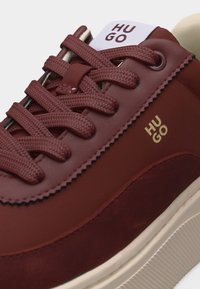 Gros plan d'une basket rouge foncé avec des lacets texturés, une semelle blanche et le logo "HUGO" sur le côté et la languette.