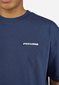 Tamsiai mėlynas medvilninis marškinėlis su apvalia kaklo iškirpte, ant priekio turi baltą siuvinėtą "PEGADOR" logotipą. Minkšta tekstūra, trumpomis rankovėmis.