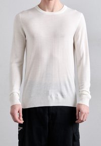 Mann in einem taillierten weißen Langarm-Pullover mit Rundhalsausschnitt und schwarzen Cargo-Hosen, der vor einem schlichten Hintergrund steht.