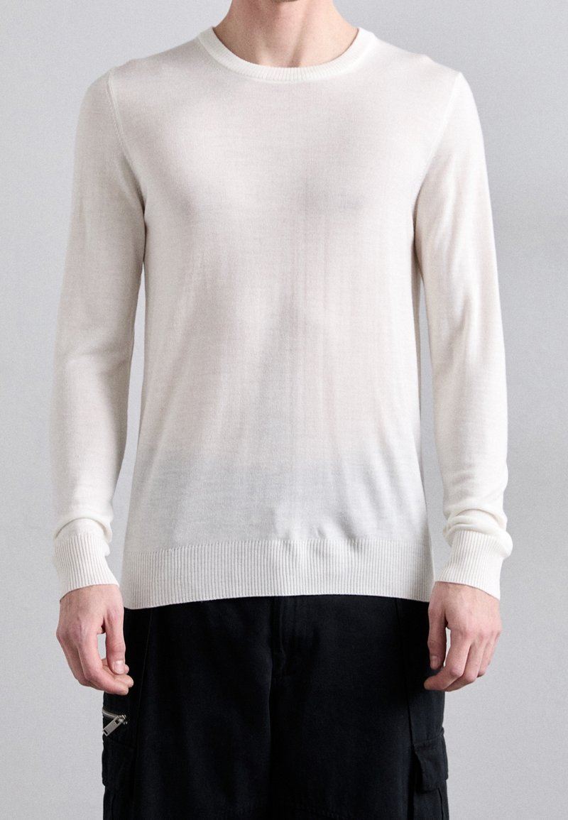 Mann in einem taillierten weißen Langarm-Pullover mit Rundhalsausschnitt und schwarzen Cargo-Hosen, der vor einem schlichten Hintergrund steht.