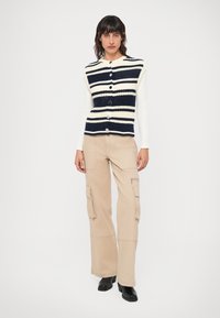 Colete de malha sem mangas apresenta riscas horizontais em azul-marinho e branco, com fecho de botões, combinado com calças cargo bege e botas de salto pretas.