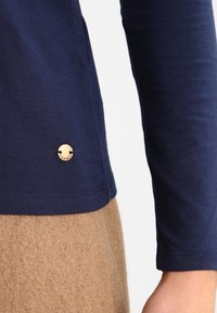 Granatowa koszula z długim rękawem, wykonana z miękkiego materiału, z zaokrąglonym dołem i małym złotym logo na dolnym prawym rogu.