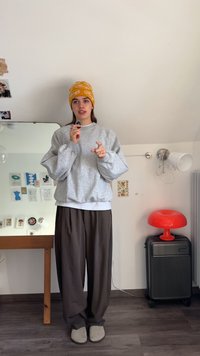 Felpa grigia con spalle scese, abbinata a pantaloni marroni a gamba larga. Indossa un berretto giallo, tiene in mano un piccolo oggetto. La stanza presenta uno specchio e una lampada rossa.