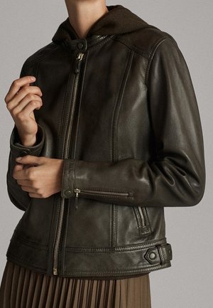Person wearing a dark brown leather jacket with front zipper, zippered cuffs, button details, over a brown pleated skirt.