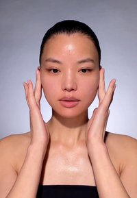 Frau mit glatter, hydratisierter Haut, die ein schwarzes, trägerloses Oberteil trägt. Ihre Hände berühren sanft ihr Gesicht und zeigen einen natürlichen Glanz.