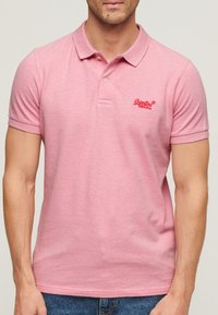Różowa koszulka polo wykonana z bawełny, z kołnierzykiem na dwa guziki, krótkimi rękawami oraz czerwonym haftowanym logo po lewej stronie na wysokości klatki piersiowej.