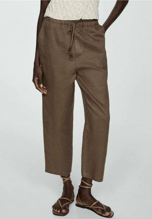 Person iført brune snorbukser, en hvid strikket top og brune flade sandaler med stropper, står med hænderne nær lommerne.