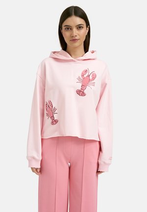 Woman wearing a light pink cropped hoodie with two pink lobster designs, paired with loose pink trousers against a plain background.