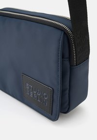 Dunkelblaue, strukturierte Nylon-Tasche in rechteckiger Form, mit einem silbernen Reißverschluss, einem schwarzen Stoffriemen und einem schwarzen, geprägten Etikett mit der Aufschrift "STUDIO BERLIN."