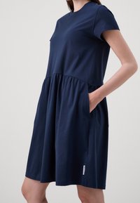 Vrouw in een marineblauw jurkje met korte mouwen en een vergaarde taille, met zijzakken, staand tegen een effen lichte achtergrond.