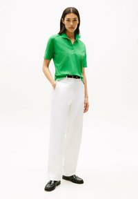 Zielona koszulka polo z krótkim rękawem, z trzema guzikami przy kołnierzyku, w zestawieniu z białymi, szerokimi spodniami i czarnymi loafersami, stojąca na neutralnym tle.