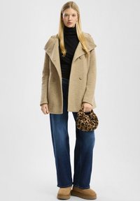 Beež villasegu mantel laia krae ja kahekordse nööpimisega, koos tumesiniste laiade pükstega ja leopardi mustriga käekotiga.