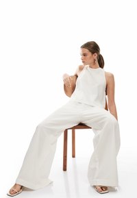 Top bianco senza maniche con collo alto, abbinato a pantaloni larghi realizzati in un tessuto morbido. Modella seduta su una sedia di legno.