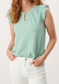 Mátově zelený top s krátkým rukávem, s výstřihem ve tvaru klíče a nařeněnými ramenními detaily, v kombinaci s bílými kalhotami. Měkká, texturovaná tkanina.