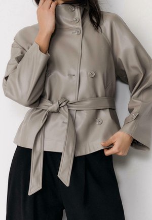 Femme portant une veste en cuir gris clair ceinturée avec de gros boutons, par-dessus un pantalon noir, une main près du col et l’autre à l’ourlet.