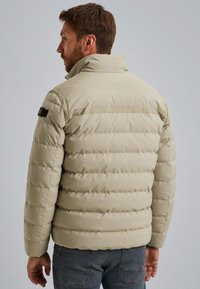 Veste en duvet beige avec un motif de coutures horizontales, col montant et accent logo noir sur l'épaule, vue de dos.