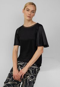 Czarna satynowa bluzka z krótkimi, rozszerzanymi rękawami, okrągłym dekoltem z metaliczną lamówką, zestawiona z wzorzystymi czarno-białymi spodniami.