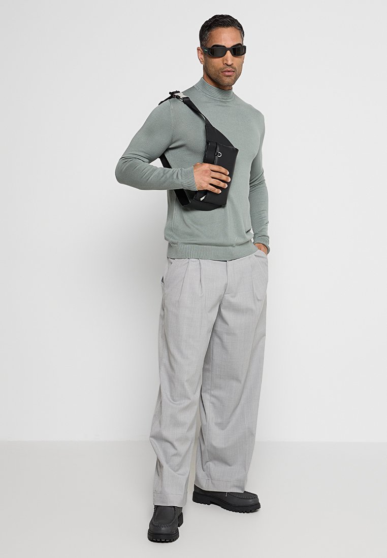 Grau gestrickter Rollkragenpullover, hellgraue plissierte Hosen, schwarze Umhängetasche und schwarze Sonnenbrille. Robuste schwarze Schuhe mit strukturierten Sohlen.