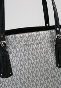 Sac à main Michael Kors argenté avec des sangles en cuir noir, présentant un motif répétitif du logo MK et une plaque métallique avec le nom de la marque à l'avant.