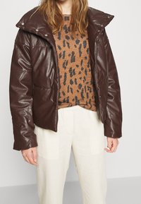 Braune Kunstleder-Pufferjacke mit hohem Kragen, Druckknöpfen, getragen über einem tan- und schwarz-gefleckten Pullover und hellen Hosen.