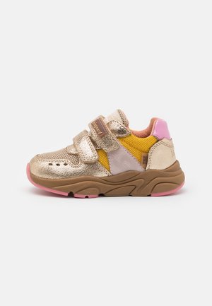 Gouden en roze sneakers met gele mesh-accenten, voorzien van twee verstelbare klittenbandbanden en een grove, gestructureerde bruine zool.