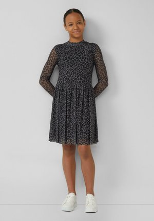 Schwarzes Leopardenmuster-Kleid mit langen, transparenten Netzärmeln und einem gerafften Rock; kombiniert mit weißen Sneakers. Glatte Stoffstruktur.