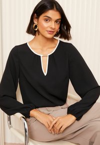 Blusa nera con rifiniture bianche, scollatura a chiave e maniche lunghe a sbuffo. Modella che indossa un bracciale semplice. Tessuto liscio con una leggera lucentezza.