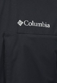 Λογότυπο Columbia σε λευκό χρώμα σε μαύρο ύφασμα πάνω από μια οριζόντια τσέπη με ορατή ραφή και φερμουάρ στην αριστερή πλευρά.