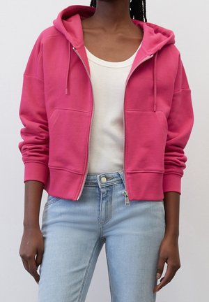 Persoon die een felroze hoodie met rits draagt over een wit tanktopje, gecombineerd met lichtblauwe jeans met een hoge taille.