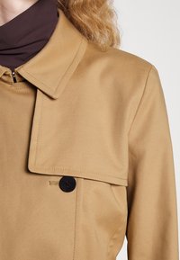 Trench-coat marron clair avec un large col et un bouton noir, porté sur un pull à col roulé marron foncé.