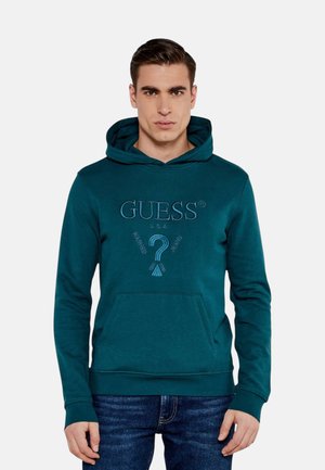 Młody mężczyzna w zielononiebieskiej bluzie Guess z kapturem z przednią kieszenią i wyszywanym logo, w połączeniu z niebieskimi dżinsami, stojący na jednolitym tle.