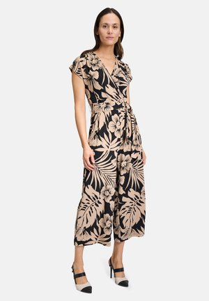 Combinaison en noir avec un motif floral beige, manches courtes, taille cintrée et jambes larges. Associée à des chaussures à talons hauts.